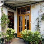 Cozy café entrance with wooden door surrounded by lush green plants and potted flowers.